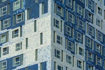 Close-up view of a modern building with mosaic tiles in varying shades of blue and white. The geometric pattern creates a textured, dynamic facade with many windows.