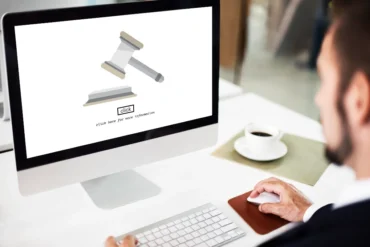 A man in a suit uses a computer mouse at a desk. The monitor displays a gavel and a button labeled "Click for more information." A coffee cup sits nearby.