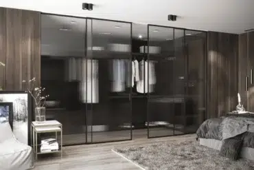 Modern bedroom with a sleek design, featuring glass-door wardrobes, wood panel walls, a gray bed, plush rug, decorative art, and serene lighting.