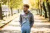 A man stands on a leaf-strewn path lined with autumn trees, wearing a gray "Nirvana" hoodie and denim overalls, exuding a calm, reflective mood.