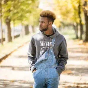 A man stands on a leaf-strewn path lined with autumn trees, wearing a gray "Nirvana" hoodie and denim overalls, exuding a calm, reflective mood.