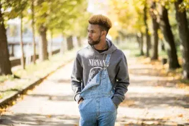 A man stands on a leaf-strewn path lined with autumn trees, wearing a gray "Nirvana" hoodie and denim overalls, exuding a calm, reflective mood.