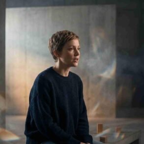 A person with short hair sits on a bench in a dimly lit room with textured walls. Soft light and shadows create a contemplative, serene atmosphere.