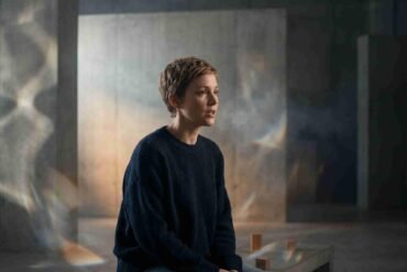 A person with short hair sits on a bench in a dimly lit room with textured walls. Soft light and shadows create a contemplative, serene atmosphere.