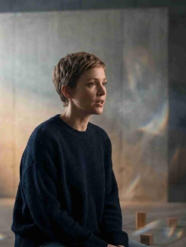 A person with short hair sits on a bench in a dimly lit room with textured walls. Soft light and shadows create a contemplative, serene atmosphere.