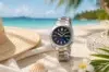 A silver watch with a blue face is elegantly placed on a sandy beach beside a straw hat, shell, and white flower. Clear blue sea and palm trees are in the background, evoking relaxation and luxury.