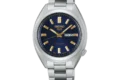 A silver metal Seiko watch with a blue dial, gold hour markers, and hands, displaying the day and date. The tone is elegant and sophisticated.