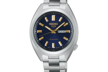 A silver metal Seiko watch with a blue dial, gold hour markers, and hands, displaying the day and date. The tone is elegant and sophisticated.