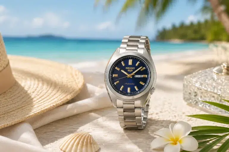 A silver watch with a blue face is elegantly placed on a sandy beach beside a straw hat, shell, and white flower. Clear blue sea and palm trees are in the background, evoking relaxation and luxury.
