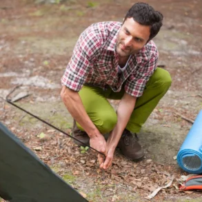 Man assembling a tent in a forest, wearing a plaid shirt and green pants. A rolled blue sleeping mat and orange backpack are nearby on the ground.
