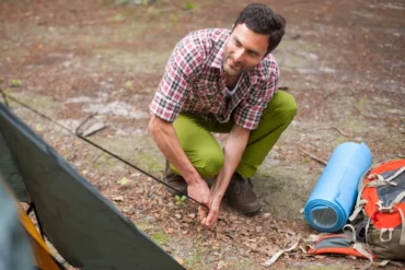 Man assembling a tent in a forest, wearing a plaid shirt and green pants. A rolled blue sleeping mat and orange backpack are nearby on the ground.