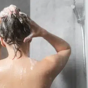 Person washing hair in a modern shower. Bubbles cover wet hair, while water droplets glisten on skin. The mood is calm and refreshing.