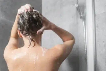 Person washing hair in a modern shower. Bubbles cover wet hair, while water droplets glisten on skin. The mood is calm and refreshing.