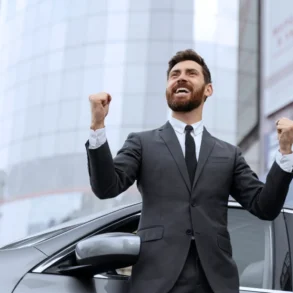 A man in a suit stands beside a car, fists raised in triumph. He smiles broadly, expressing excitement. A modern building looms in the background.