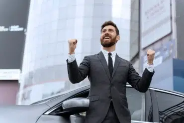 A man in a suit stands beside a car, fists raised in triumph. He smiles broadly, expressing excitement. A modern building looms in the background.