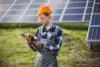 A worker in a plaid shirt and orange hard hat examines a tablet while standing in front of a solar panel array on a sunny day, focusing intently.