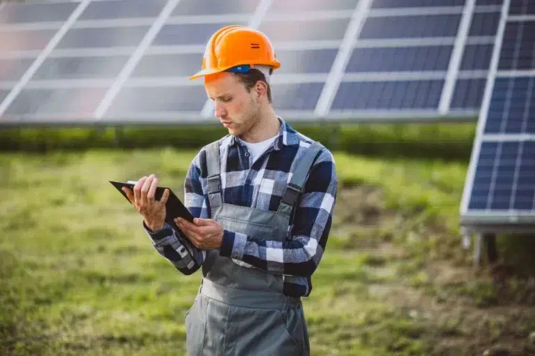 A worker in a plaid shirt and orange hard hat examines a tablet while standing in front of a solar panel array on a sunny day, focusing intently.