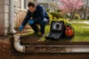 A man inspects an outdoor pipe with a camera cable, shown on a screen. Scene reveals underground pipe system, surrounded by grass and spring trees.