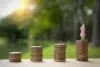 Stacks of coins increase in height from left to right, with a small figurine of a woman in a pink dress standing on the tallest stack. Sunlight filters through greenery in the background, creating a hopeful tone.