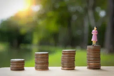 Stacks of coins increase in height from left to right, with a small figurine of a woman in a pink dress standing on the tallest stack. Sunlight filters through greenery in the background, creating a hopeful tone.