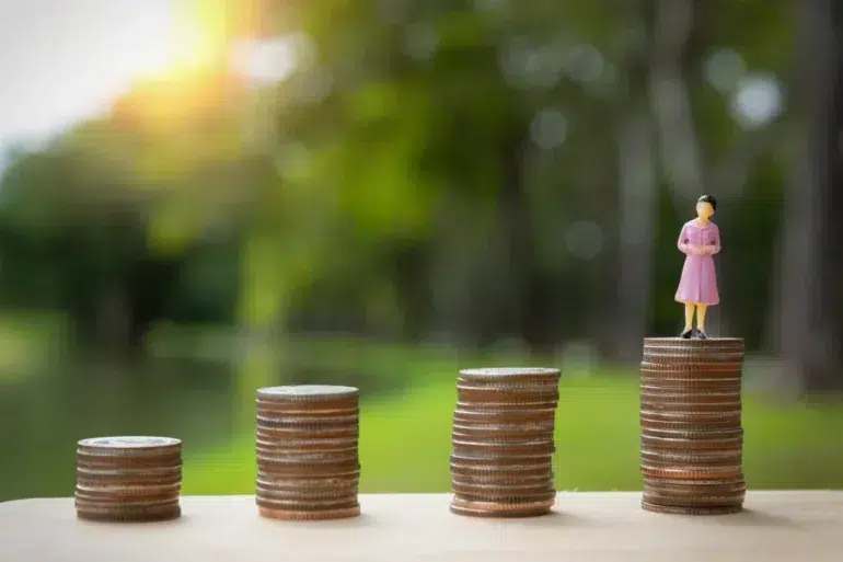 Stacks of coins increase in height from left to right, with a small figurine of a woman in a pink dress standing on the tallest stack. Sunlight filters through greenery in the background, creating a hopeful tone.