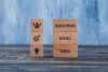 Wooden blocks on a table display motivational concepts. One stack shows "SUCCESS," "GOAL," "IDEA"; the other has icons for achievement and creativity.