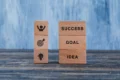 Wooden blocks on a table display motivational concepts. One stack shows "SUCCESS," "GOAL," "IDEA"; the other has icons for achievement and creativity.