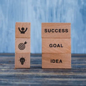 Wooden blocks on a table display motivational concepts. One stack shows "SUCCESS," "GOAL," "IDEA"; the other has icons for achievement and creativity.