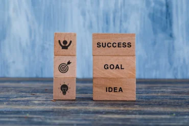 Wooden blocks on a table display motivational concepts. One stack shows "SUCCESS," "GOAL," "IDEA"; the other has icons for achievement and creativity.