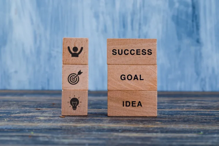 Wooden blocks on a table display motivational concepts. One stack shows "SUCCESS," "GOAL," "IDEA"; the other has icons for achievement and creativity.