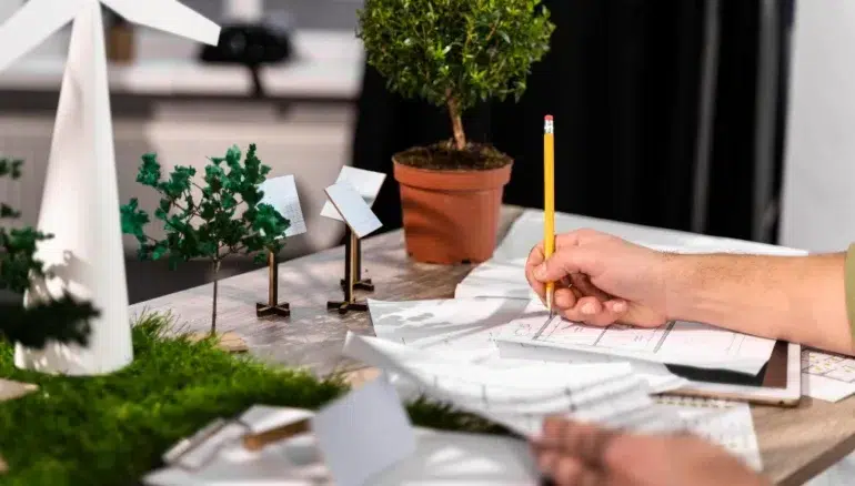 A person sketches designs on paper at a desk with model trees, wind turbines, and solar panels, conveying a focus on sustainable energy and planning.