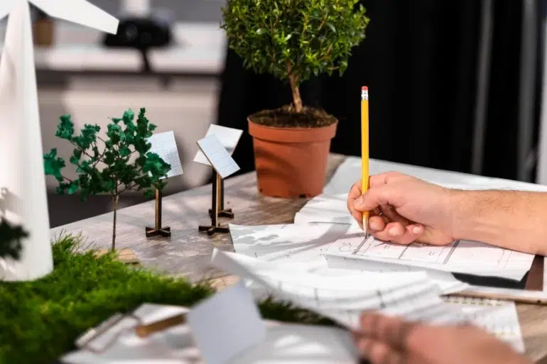A person sketches designs on paper at a desk with model trees, wind turbines, and solar panels, conveying a focus on sustainable energy and planning.