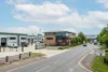 An elevated view of a paved entrance into Suttons Business Park, showing industrial buildings on the left with EV chargers and a forklift, a three-story office building with a Costa Coffee at its base, and a road leading towards a large 'Suttons Business Park' sign in the distance under a partly cloudy sky.