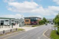 An elevated view of a paved entrance into Suttons Business Park, showing industrial buildings on the left with EV chargers and a forklift, a three-story office building with a Costa Coffee at its base, and a road leading towards a large 'Suttons Business Park' sign in the distance under a partly cloudy sky.