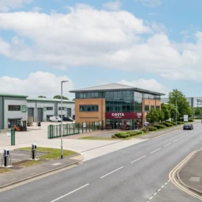 An elevated view of a paved entrance into Suttons Business Park, showing industrial buildings on the left with EV chargers and a forklift, a three-story office building with a Costa Coffee at its base, and a road leading towards a large 'Suttons Business Park' sign in the distance under a partly cloudy sky.
