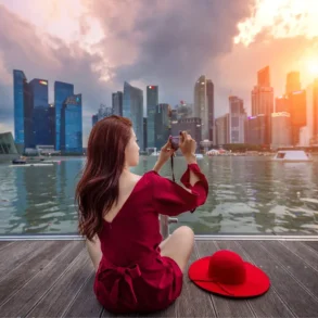 A woman in a red dress sits on a wooden deck capturing a sunset cityscape across water, with tall buildings and dramatic clouds in the background.