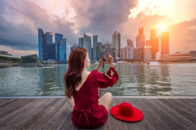 A woman in a red dress sits on a wooden deck capturing a sunset cityscape across water, with tall buildings and dramatic clouds in the background.