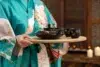 A person in a floral kimono holds a wooden tray with a black teapot and matching cups. The background is softly lit with glowing candles, creating a serene ambiance.