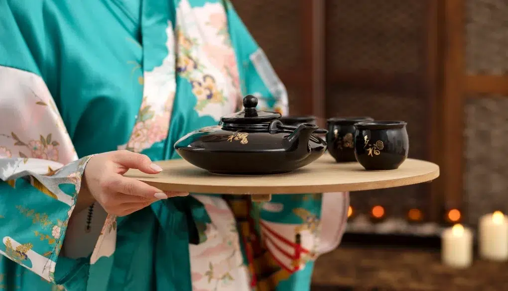 A person in a floral kimono holds a wooden tray with a black teapot and matching cups. The background is softly lit with glowing candles, creating a serene ambiance.