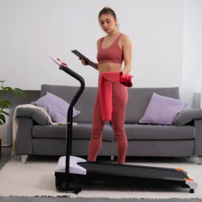 A woman in workout attire stands on a treadmill holding a tablet and red towel in a cozy living room with a gray sofa and plants, appearing focused.