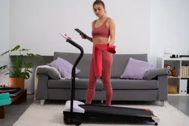 A woman in workout attire stands on a treadmill holding a tablet and red towel in a cozy living room with a gray sofa and plants, appearing focused.
