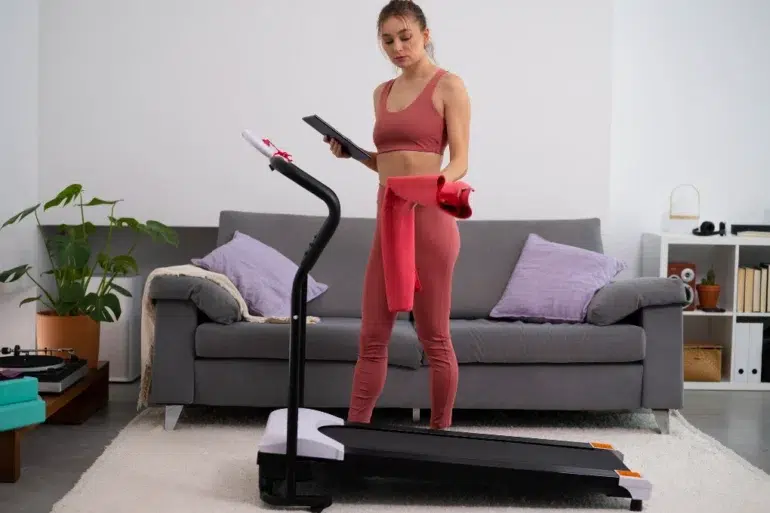 A woman in workout attire stands on a treadmill holding a tablet and red towel in a cozy living room with a gray sofa and plants, appearing focused.