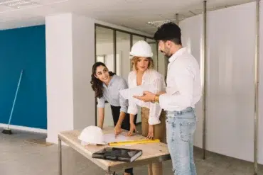 Three people in an office, wearing casual clothes and a hard hat, gather around a table with blueprints. They appear to be discussing a project.