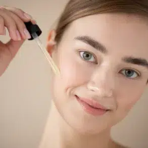 A woman with a serene smile applies facial serum with a dropper on her cheek. The background is neutral, highlighting her fresh, natural look.