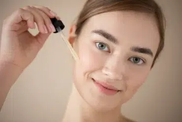 A woman with a serene smile applies facial serum with a dropper on her cheek. The background is neutral, highlighting her fresh, natural look.