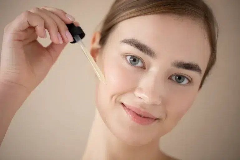 A woman with a serene smile applies facial serum with a dropper on her cheek. The background is neutral, highlighting her fresh, natural look.
