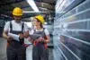 Two factory workers wearing yellow hard hats and gray overalls smile while reviewing documents on a clipboard and tablet in an industrial setting.