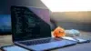 A laptop displaying code is on a wooden desk with a plush orange toy, white mouse, glasses, and notepad. Sunlight filters through a slanted window.