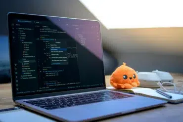 A laptop displaying code is on a wooden desk with a plush orange toy, white mouse, glasses, and notepad. Sunlight filters through a slanted window.