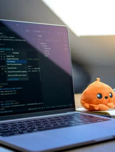 A laptop displaying code is on a wooden desk with a plush orange toy, white mouse, glasses, and notepad. Sunlight filters through a slanted window.